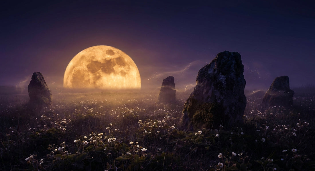 A glowing amber full moon rising low over a twilight meadow with wild blossoms and ancient standing stones silhouetted against a deep indigo sky.