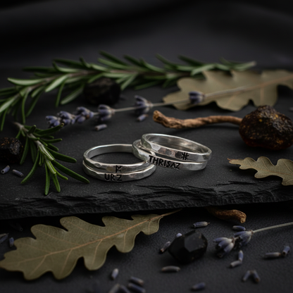Two slim hammered sterling silver rune stacking bands with Elder Futhark rune stamps, styled on dark slate with rosemary, dried oak leaves and lavender, moody talismanic jewelry product photography